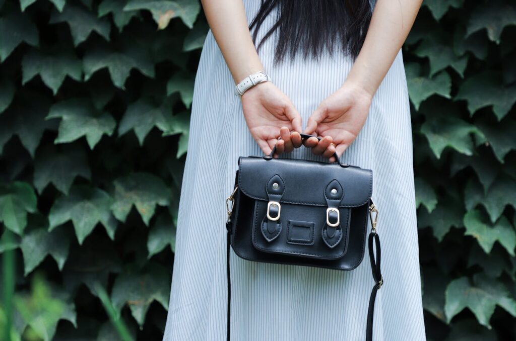 A woman in a striped dress holds a black leather handbag with lush green leaves in the background.